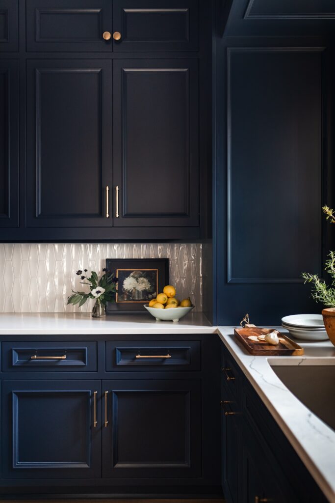 Dark blue kitchen cabinets with gold handles, white countertop, bowl of lemons, framed flower painting, cutting board, and herbs. Elegant mood.