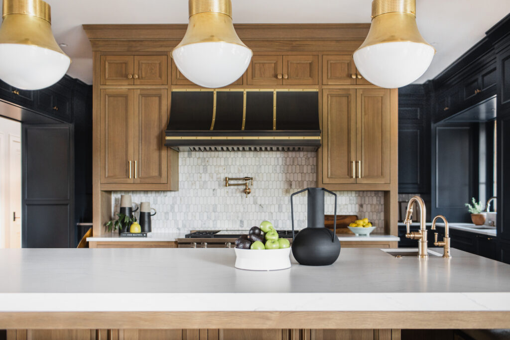 Modern kitchen with wood cabinets, black accents, and marble counter. Bowl of fruit and black pitcher on island. Brass lamps overhead.