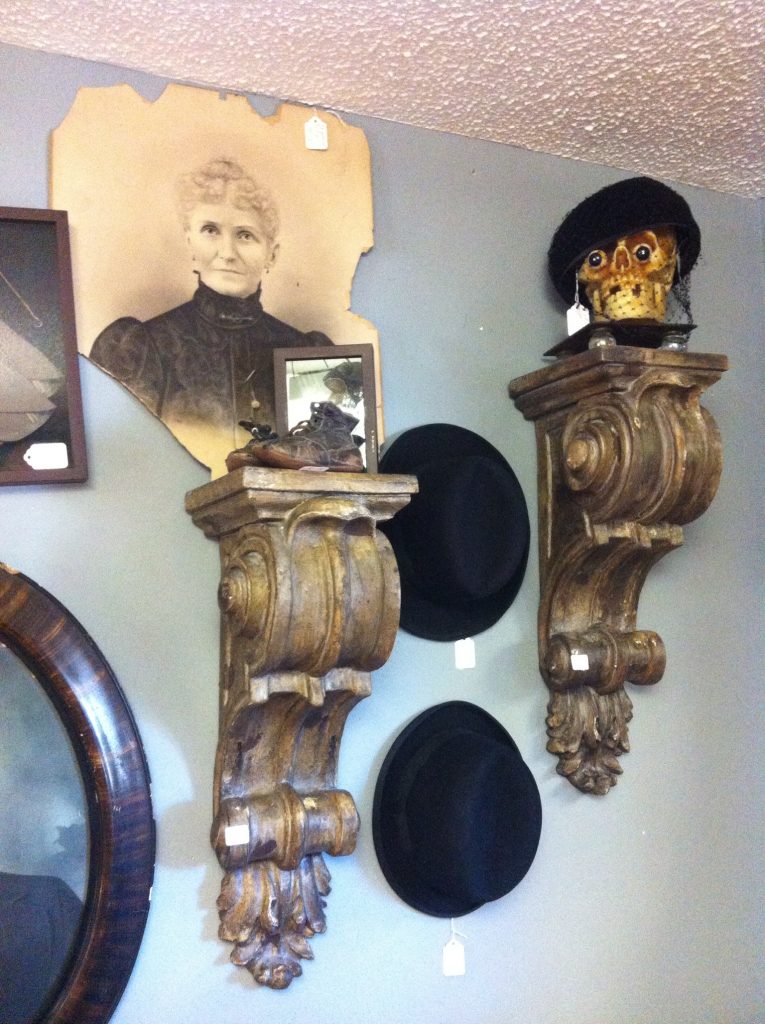Vintage wall display: framed sepia portrait, ornate shelves with hat and decorated skull, black hats below, gray textured wall.