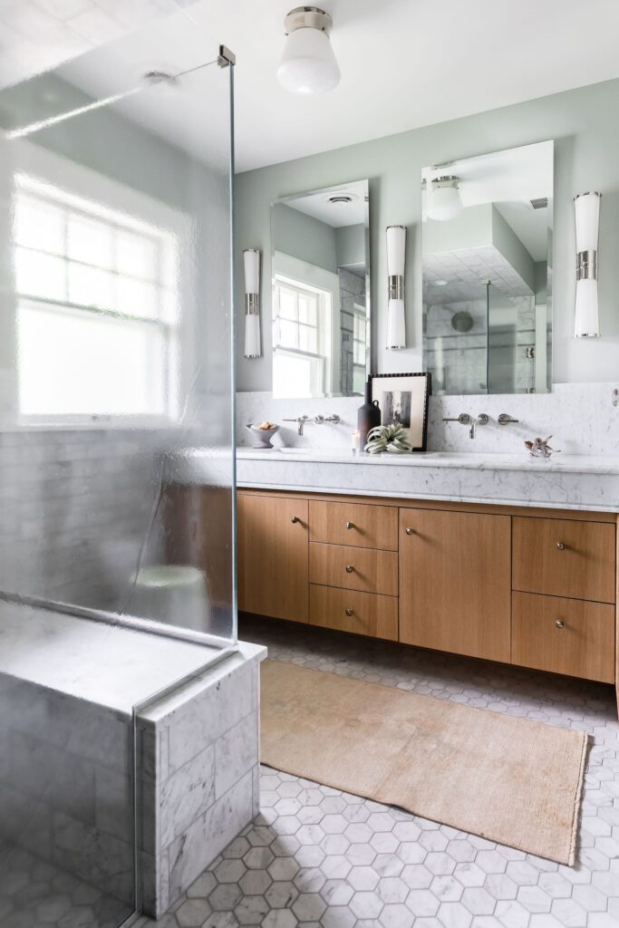Modern bathroom with wooden vanity, marble countertop, and glass shower. Hexagonal tile floor, light green walls, and soft natural light.