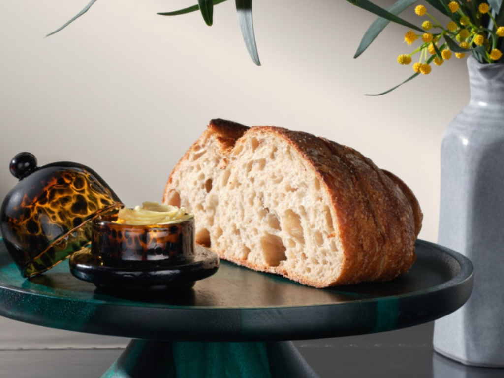 Blue Pheasant dinner display of a bread loaf on a green pedestal with butter in a tortoiseshell dish. Vase with yellow flowers in background, neutral setting.