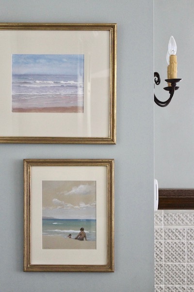 Two framed seascape paintings on a light blue wall; upper shows waves, lower depicts two people on a beach. Vintage sconce nearby.
