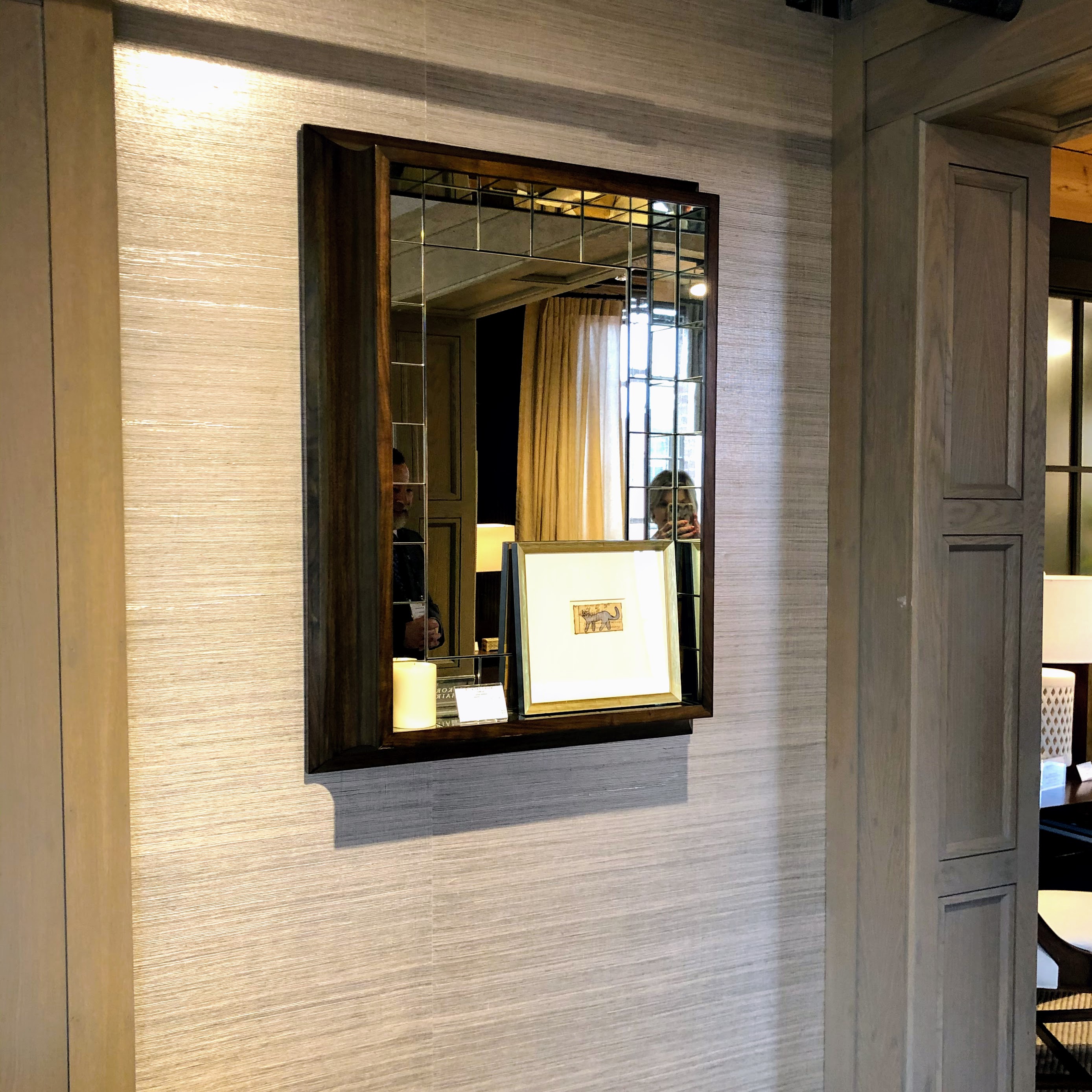 A mirror on a textured wall reflects a room with curtains and a lamp. A framed picture is in front, and two people are partially visible.
