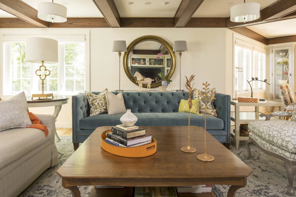 Cozy living room with blue sofa, patterned pillows, wooden coffee table with books, a decorative mirror, and large windows. Bright and inviting.