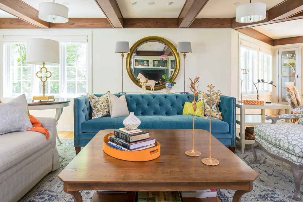 Bright living room with a blue velvet sofa, patterned pillows, wooden coffee table with books, gold decor, and a round mirror reflecting light.