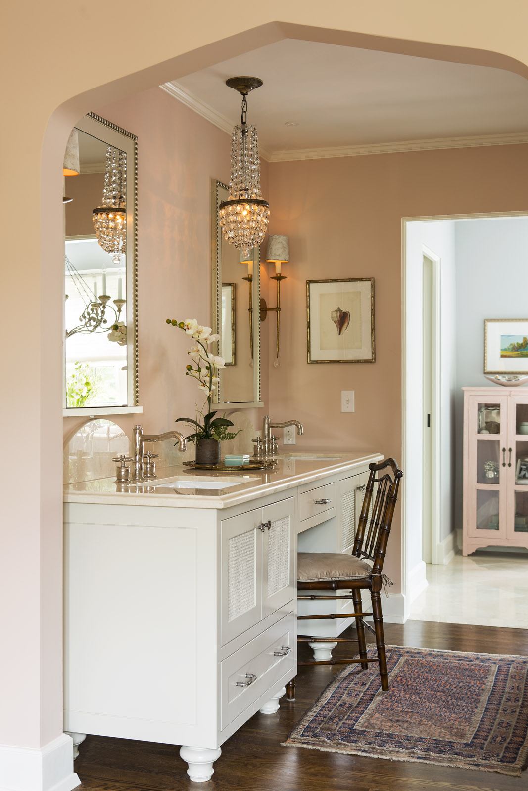 Elegant bathroom with twin sinks, crystal chandelier, and orchid. Soft pink walls, framed art, and vintage wooden chair create a cozy ambiance.