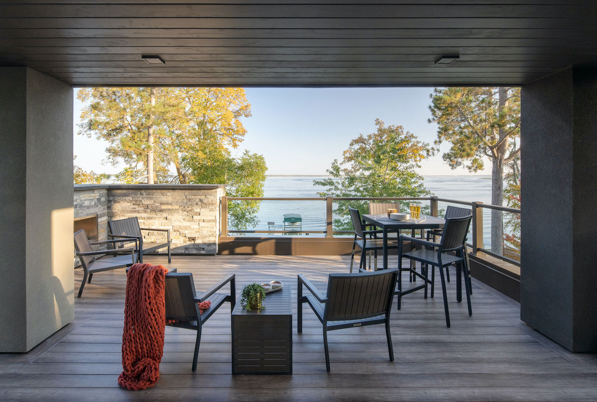 Contemporary patio with black furniture and a red blanket overlooks a serene lake. Trees and glass railing enhance the tranquil setting.
