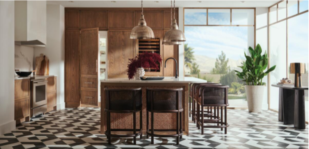 Modern kitchen with geometric floor, wooden cabinets, and island with wicker chairs. Pendant lights hang above. Large windows reveal a scenic view.