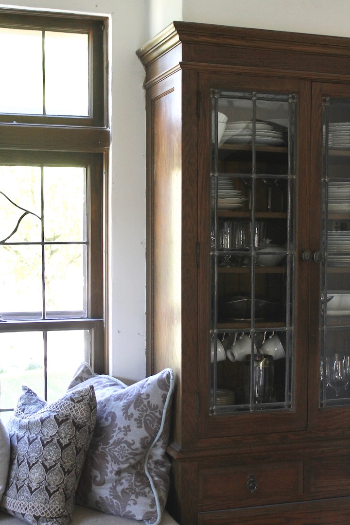 Wooden cabinet with glass doors holds plates and glasses. Patterned pillows rest on a bench by a window. Bright, cozy atmosphere.