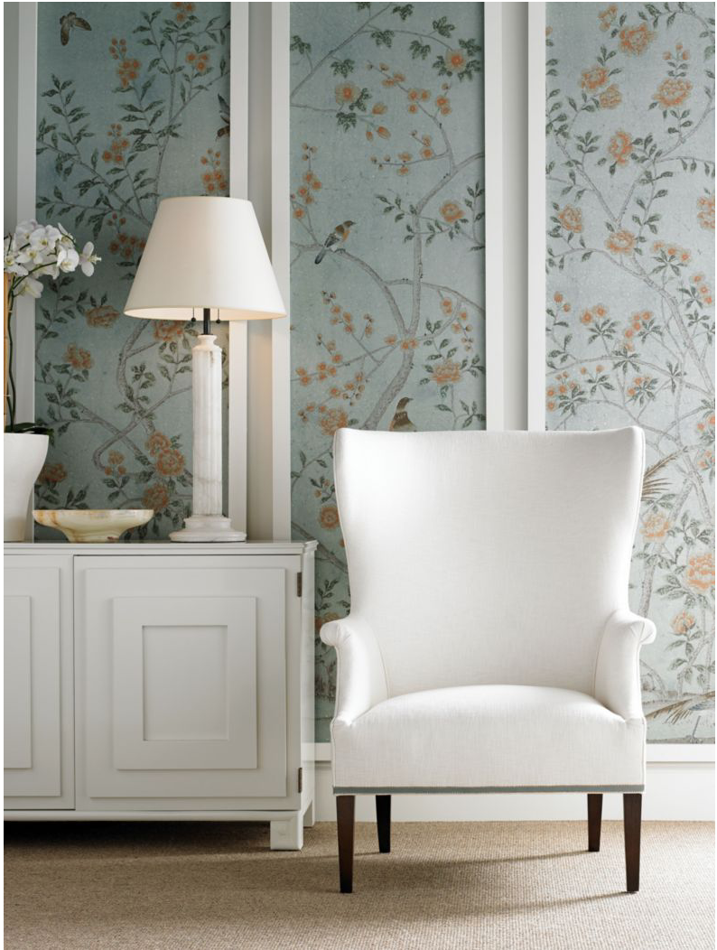 White wingback armchair by floral-patterned wallpaper. Lamp and flowers on a cabinet. Calm, elegant setting with soft colors and textures.