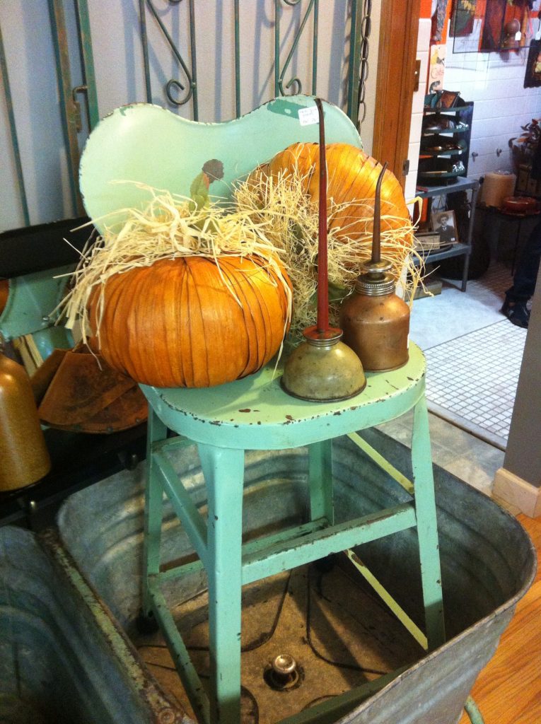 Vintage aqua chair holds pumpkins, hay, and oil cans in a rustic room. Cozy, autumnal feel with muted colors.