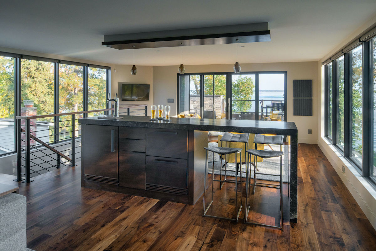 Modern kitchen with a dark island, bar stools, and glasses of beer. Large windows reveal a scenic view of trees and water. Wooden floor.