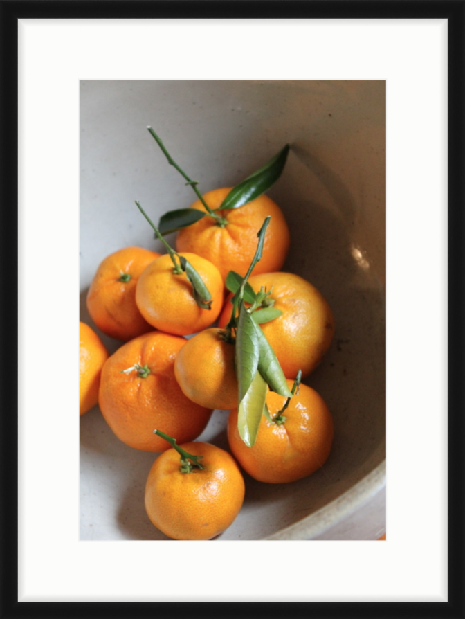 The same picture of oranges with green leaves in a bowl, framed with a wide, white mat, and thin, solid black frame. Sharp, lively mood.