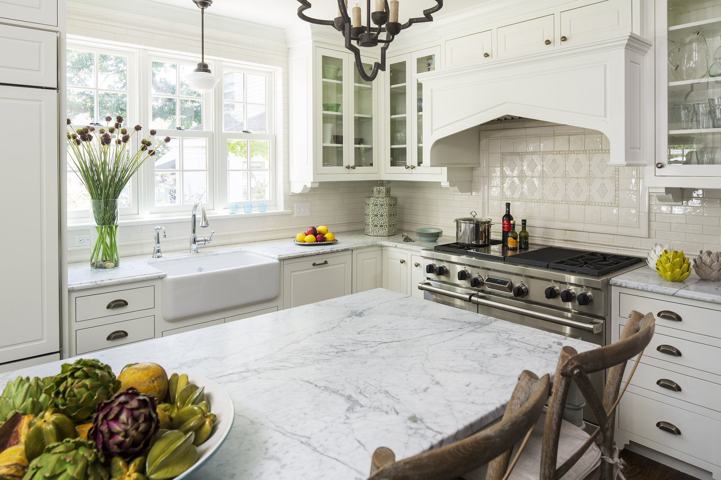 Bright kitchen with marble counters, white cabinets, and a vase of tall flowers. A bowl of vegetables, stove with pots, and fruit add color.