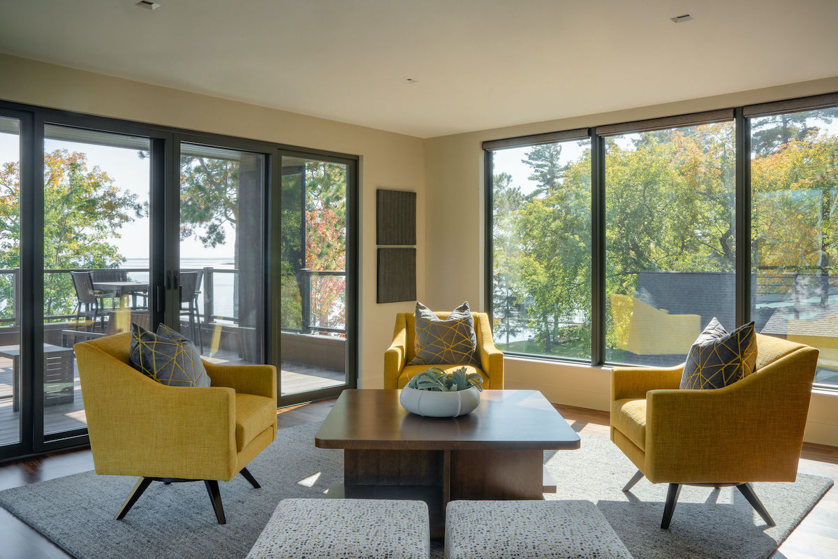 Bright living room with yellow chairs around a wooden table, large windows showing trees outside. Natural light creates a cozy ambiance.