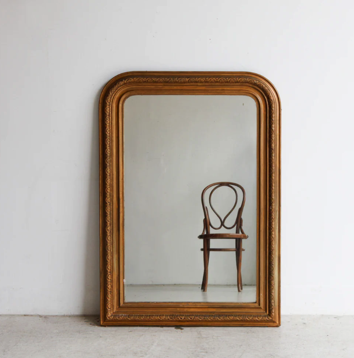 A wooden chair reflected in a large, ornate gold-framed mirror against a plain white wall, creating a minimalist, calm setting.