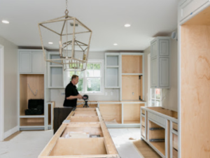 Man working on kitchen renovation with unfinished cabinets and light fixtures. Bright room with light gray walls and large windows.