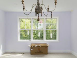 Lilac-walled room with a large window showing greenery, an open moving box in foreground, and an ornate chandelier above.