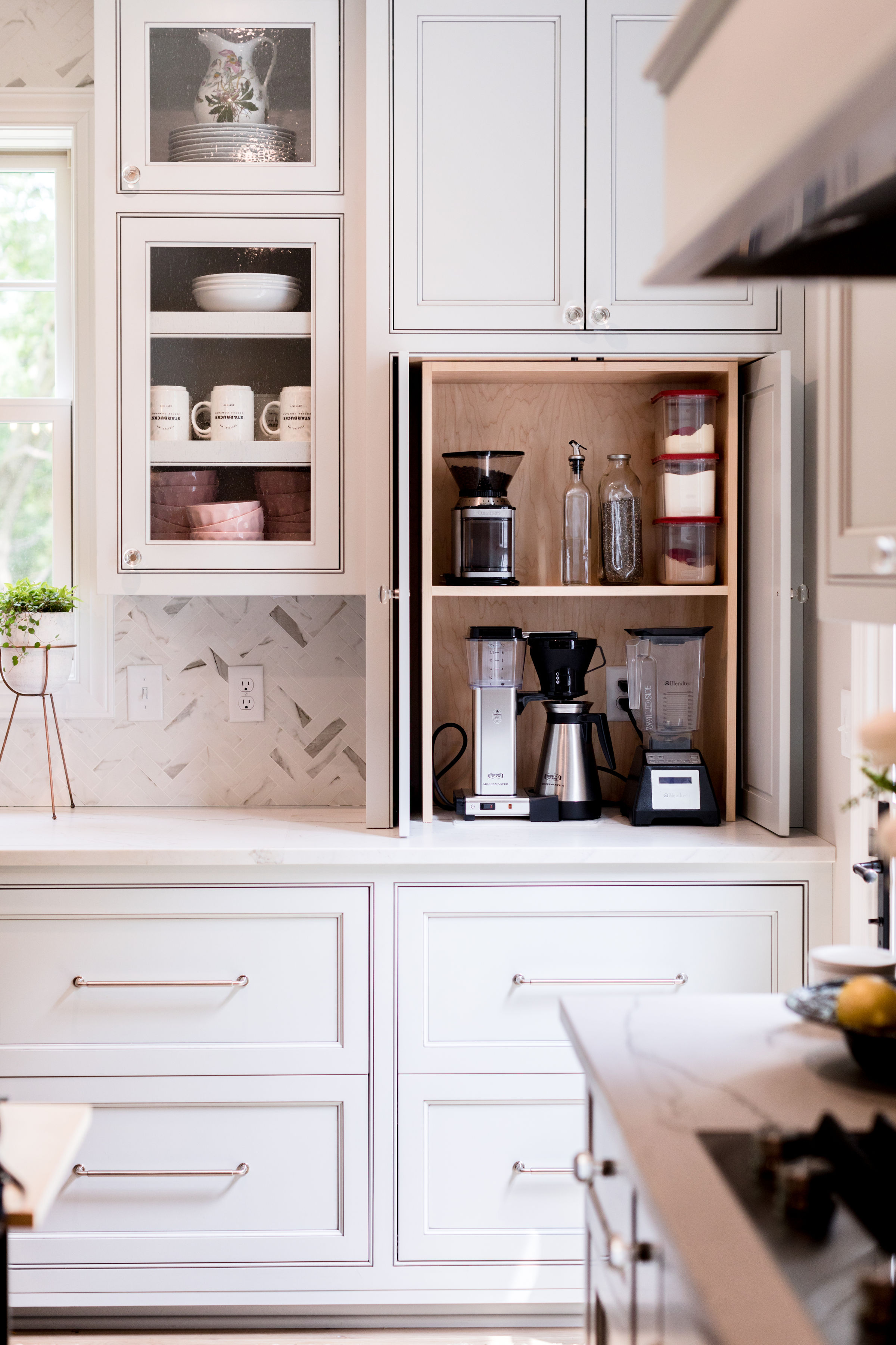 White kitchen with open cabinets showing a coffee maker, mugs, blender, and containers. Bright, tidy countertop with a plant.