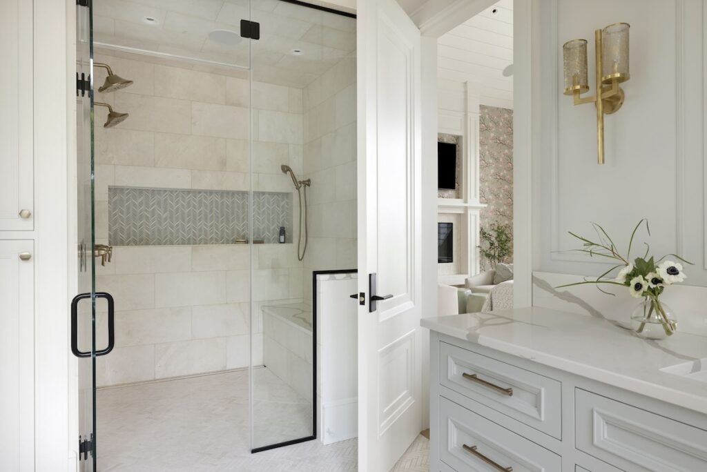 Elegant bathroom with glass shower, brass fixtures, white cabinetry, and floral wallpaper visible through an open door. Bright and serene setting.