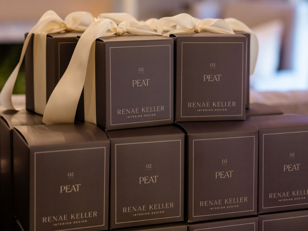 Stacked plum gift boxes tied with cream ribbons, branded "Renae Keller Interior Design" and "PEAT" text. Soft, elegant setting.