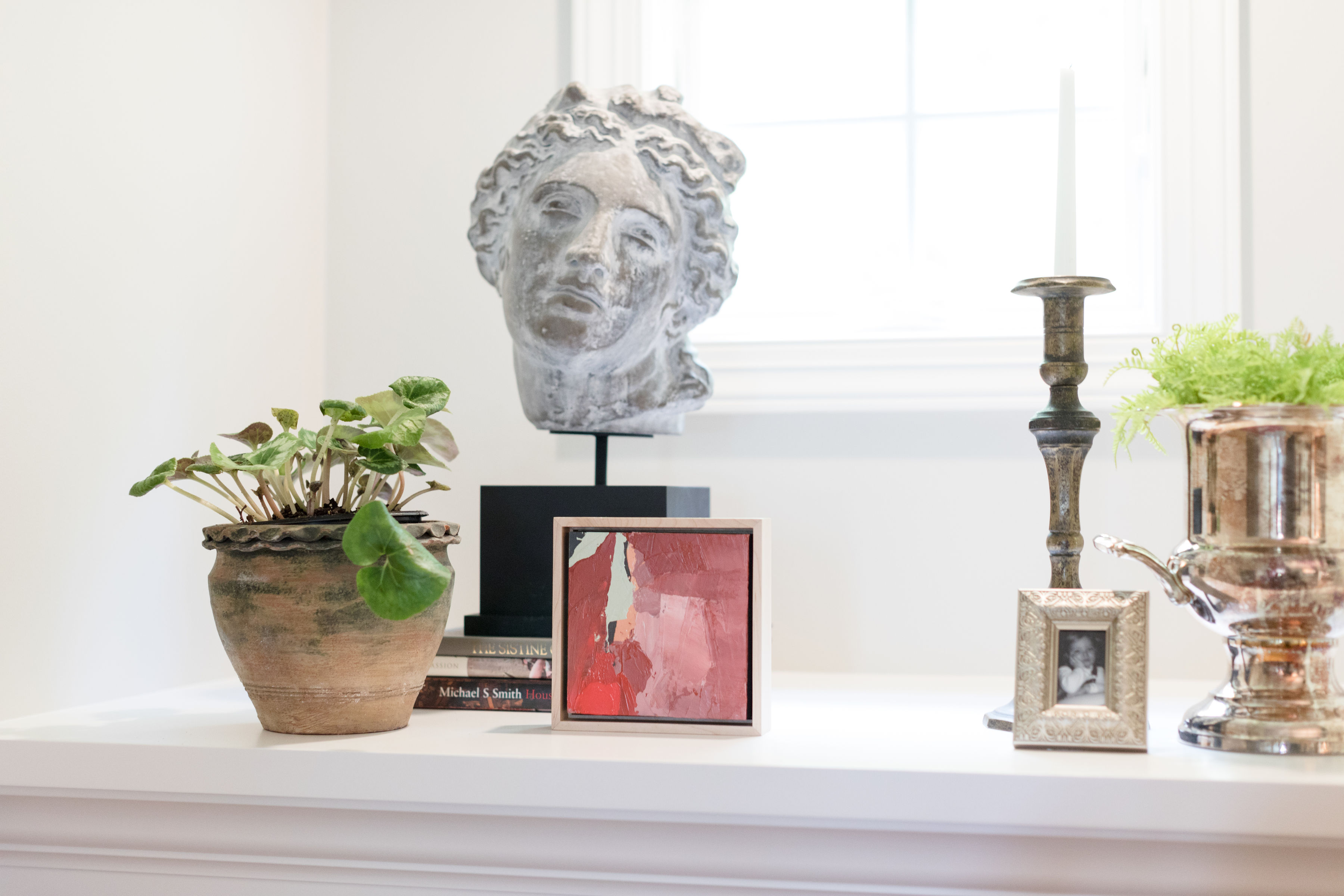 A windowsill with a plant in a pot, a stone bust, a red abstract painting, books, a candlestick, and a framed photo. Bright, minimal decor.
