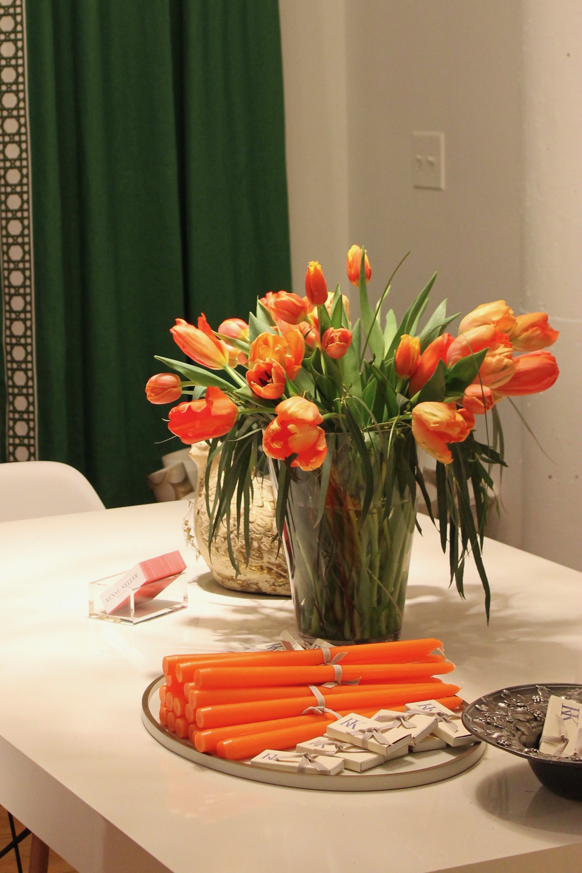 Orange tulips in a vase on a table with matching candles, small boxes, a bowl with snacks, and a patterned green curtain in the background.