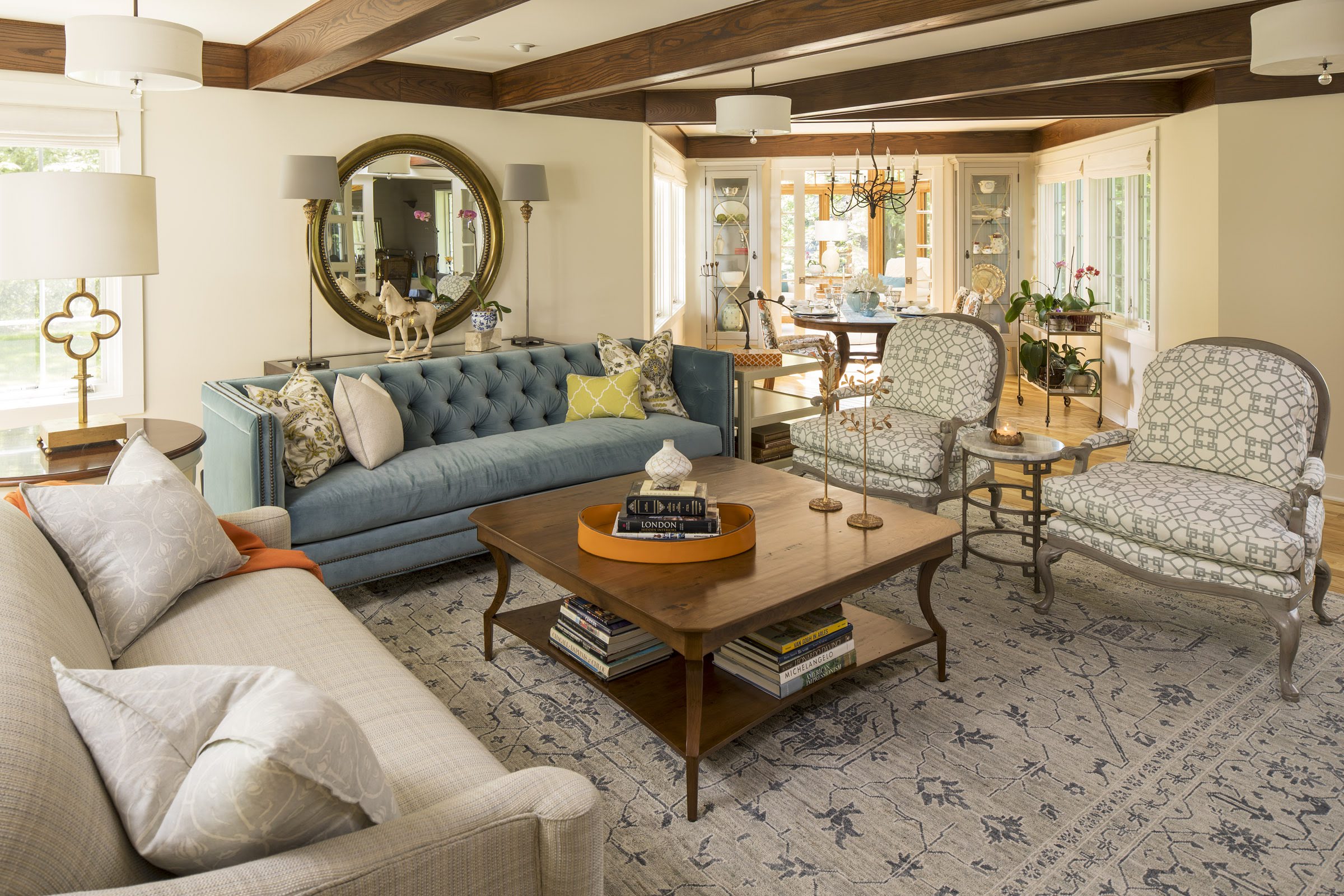 Cozy living room with blue tufted sofa, patterned chairs, and wooden coffee table. Mirror and decor accents. Bright and inviting atmosphere.