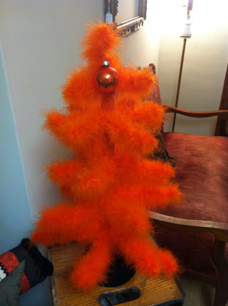 Orange fluffy feather tree with a red bauble, placed on a wooden surface in a cozy room. Background includes a red chair and a lamp.
