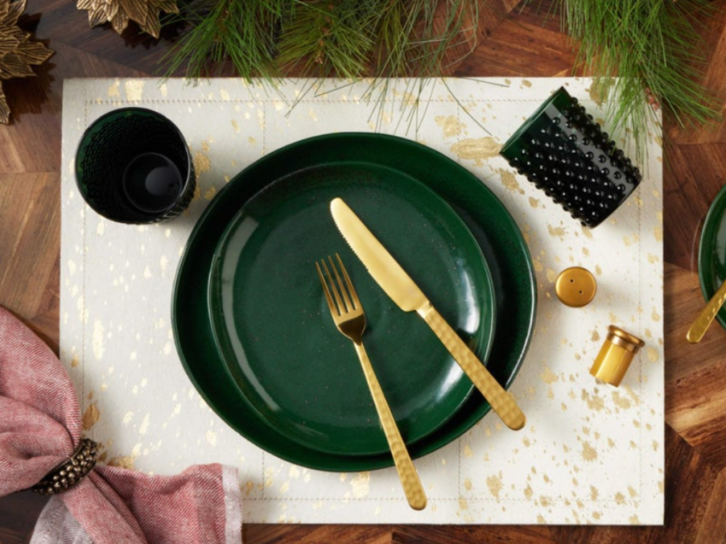Blue Pheasant green plate with gold cutlery on a speckled mat, dark green textured cup, pink napkin, pine leaves on a wooden table setting. Festive vibe.