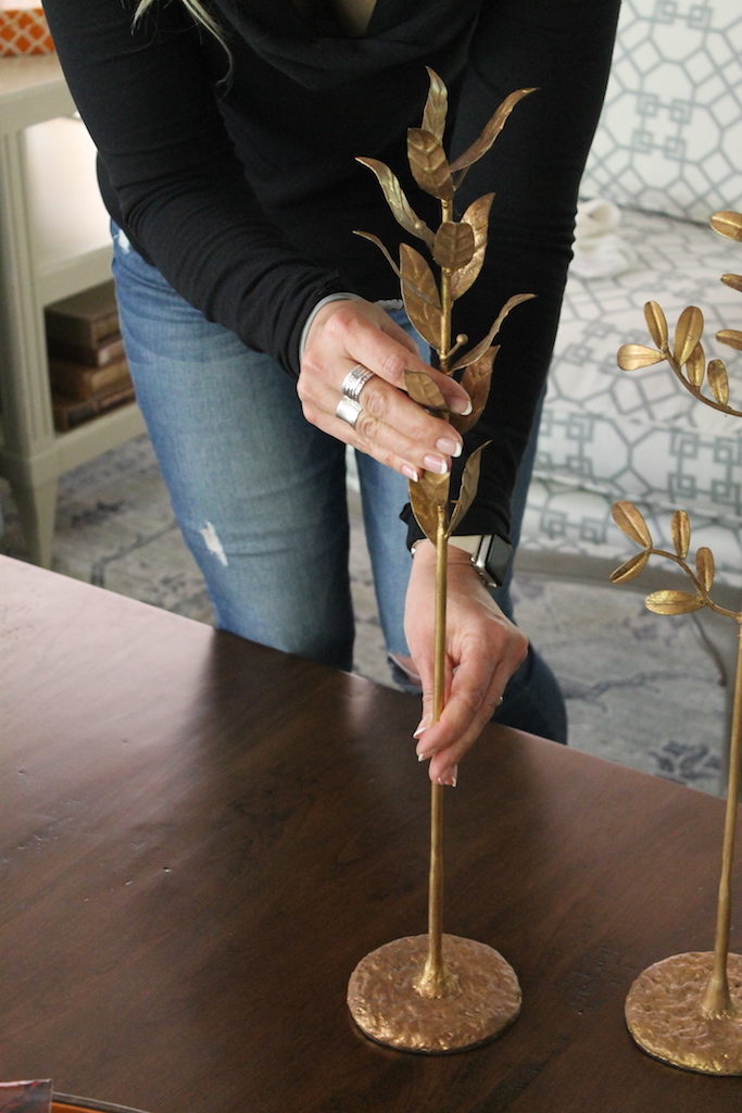 Person arranging a gold leaf sculpture on a wooden table. Background features a patterned couch and a light decor setting.