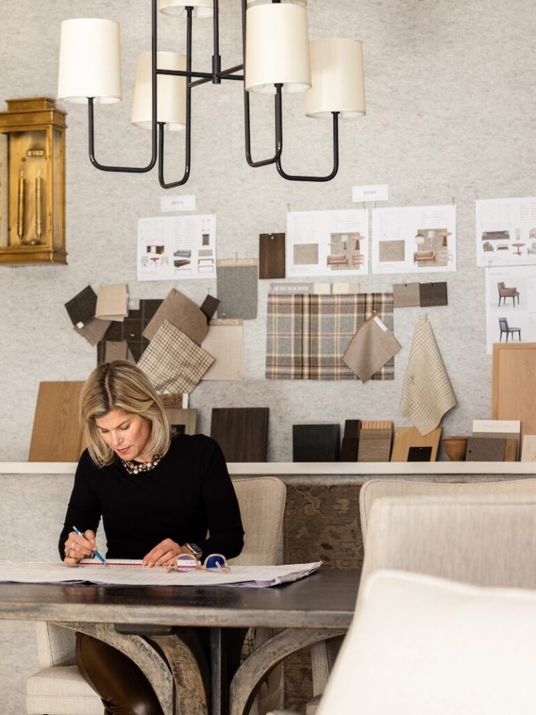 Renae works at a table with design plans. Fabric and color samples decorate the wall. Modern chandelier and neutral tones set a focused mood.