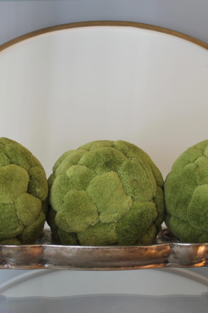 Three textured green balls on a silver tray, set against a neutral background with a curved mirror, creating a modern, serene atmosphere.