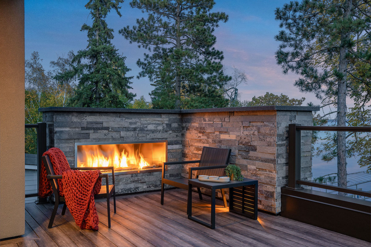 Cozy patio with fireplace, two chairs, red knit blanket, and table. Surrounded by trees at dusk, creating a warm, serene atmosphere.