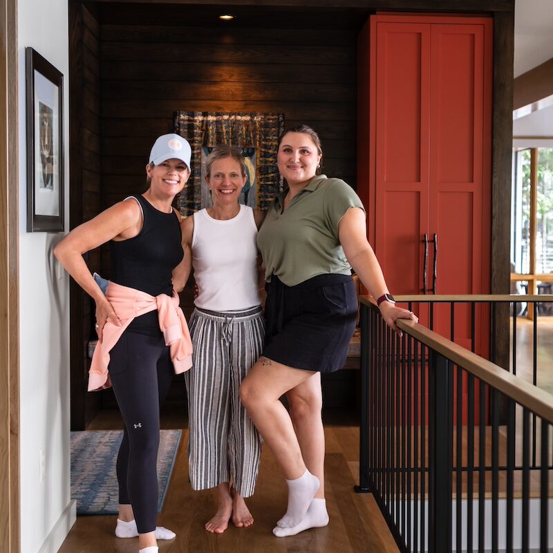 Three smiling women pose together indoors; one wears a cap, another striped pants. Behind them is a colorful rug on a dark wood wall.