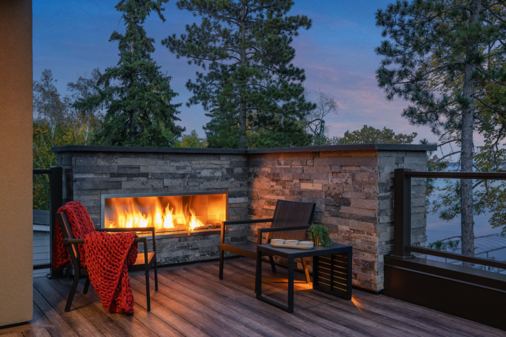Cozy patio with a modern fireplace and two chairs, one draped with a red blanket. Surrounded by trees at sunset, creating a warm atmosphere.