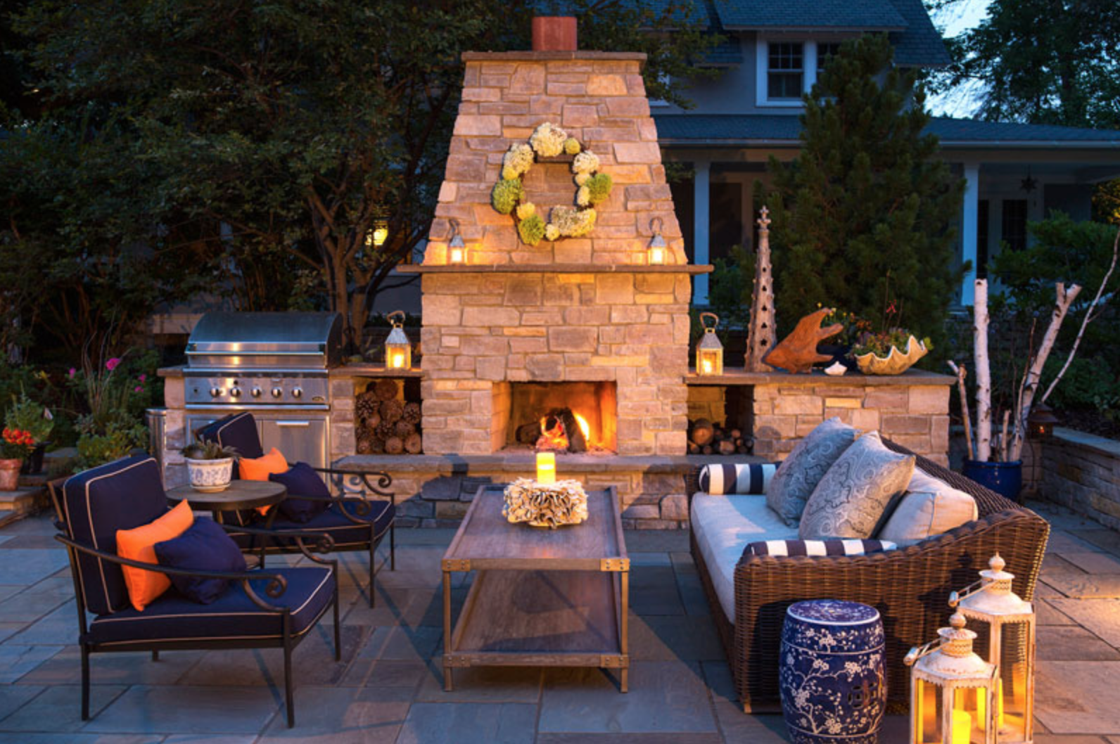 Cozy patio with a lit stone fireplace, blue and orange cushioned chairs, wicker sofa, candles, and lanterns. Twilight ambiance.