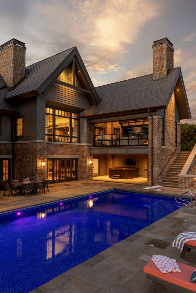 Luxurious brick house with illuminated windows beside a serene blue pool at sunset. Outdoor seating and loungers create an inviting ambiance.
