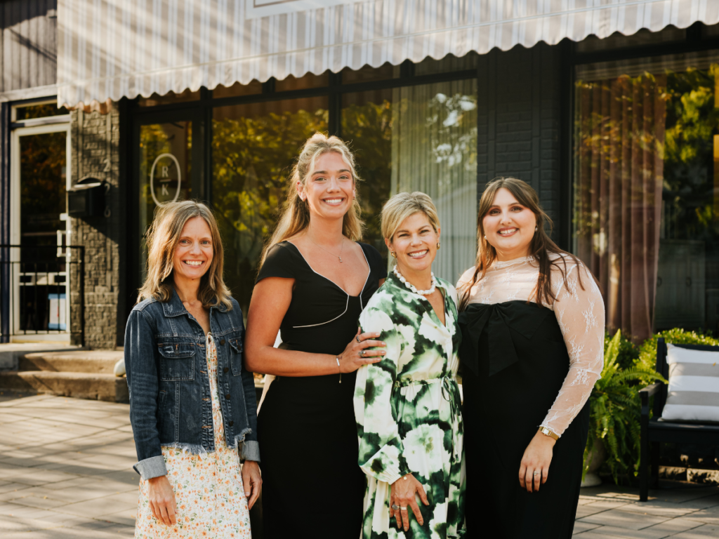 Four women smiling, standing together outside a building with a striped awning in a tree-lined neighborhood.