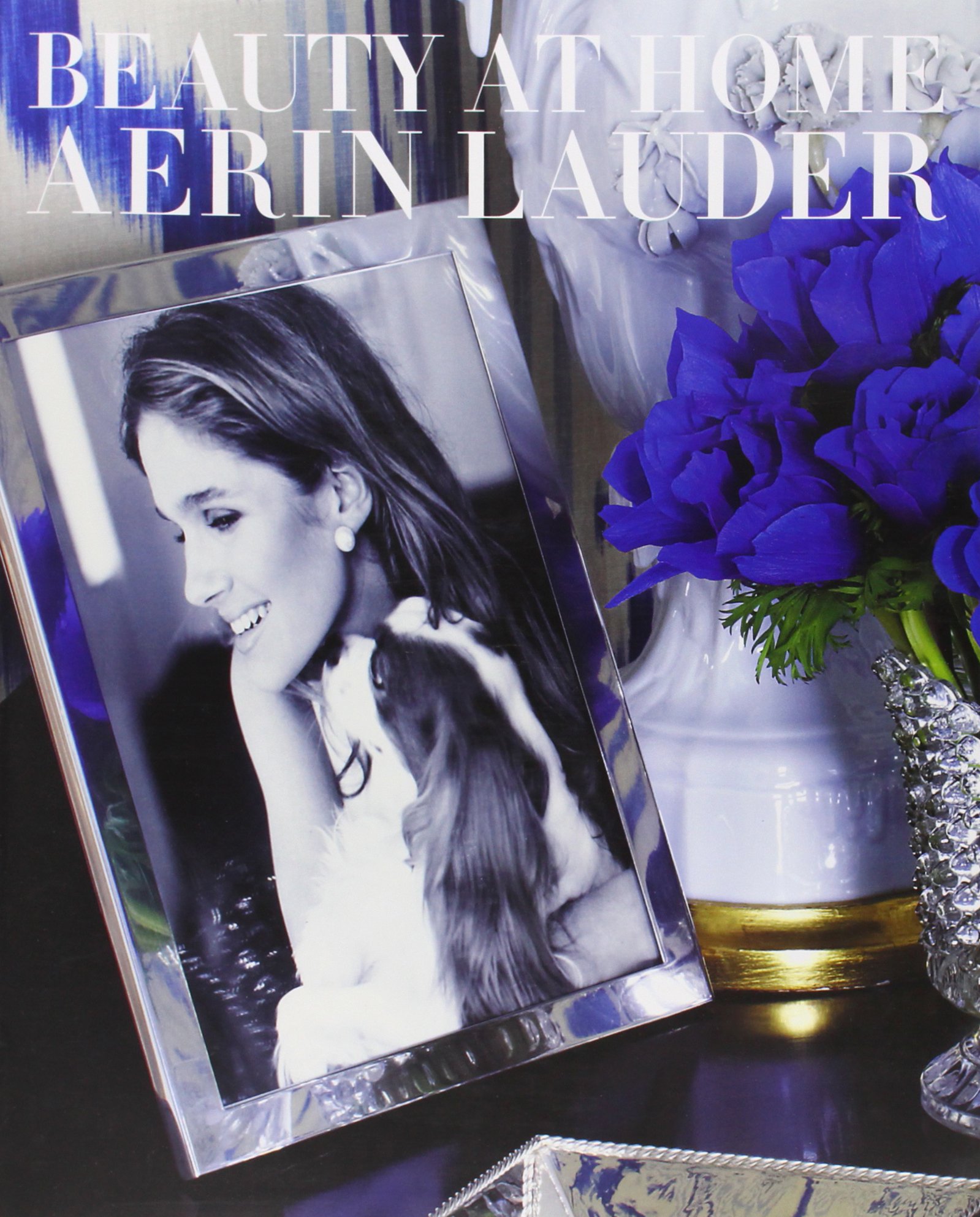 Cover of a book titled "Beauty at Home" by Aerin Lauder. Photo features a framed photo of a smiling woman with a dog, surrounded by purple flowers.