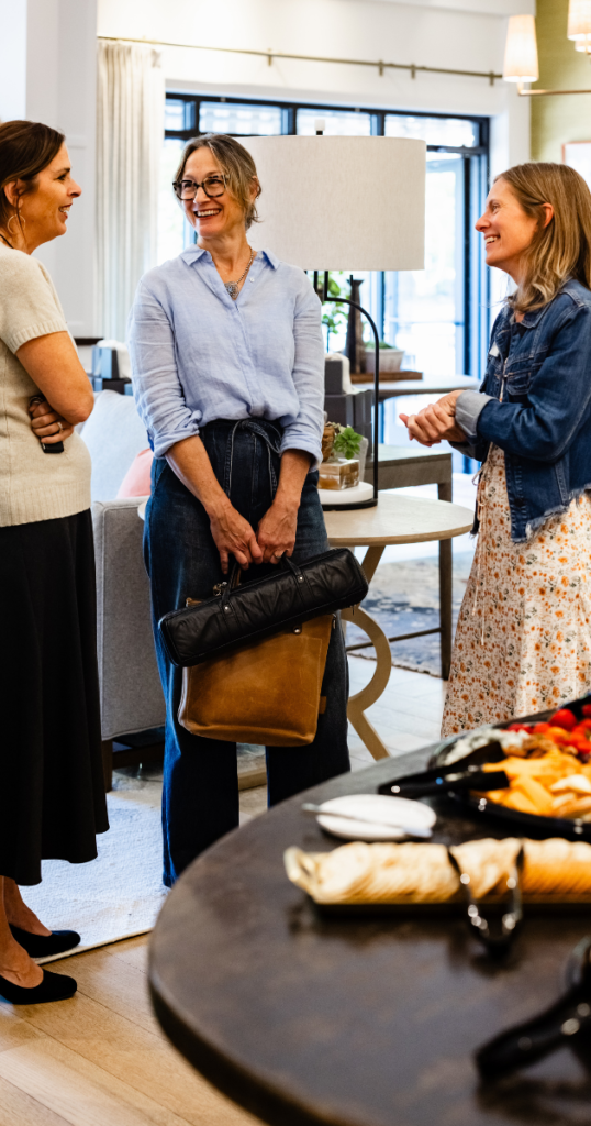 Three women chat and smile indoors near a table of appetizers. The setting is bright with a cozy, inviting atmosphere.