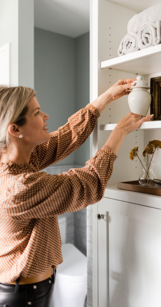 A woman in a patterned shirt places a jar on a white shelf with towels. A vase with flowers is on a cabinet in a bright bathroom.