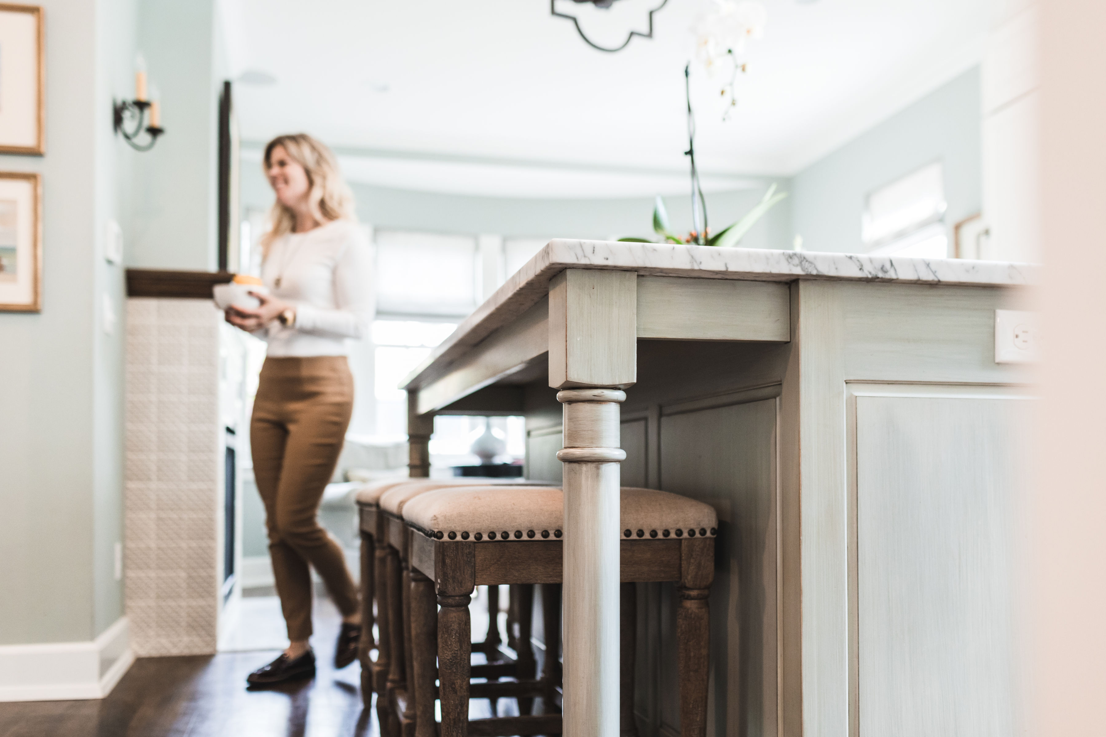 Woman holding a bowl walks in a bright kitchen with a marble island and wooden stools. Soft lighting creates a cozy atmosphere.
