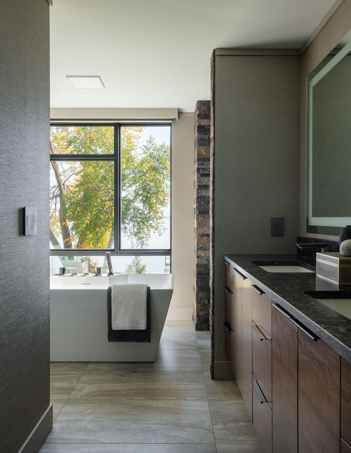 Modern bathroom with large window showing trees. White bathtub with towel, dark wood cabinets, and stone accent wall create a serene mood.