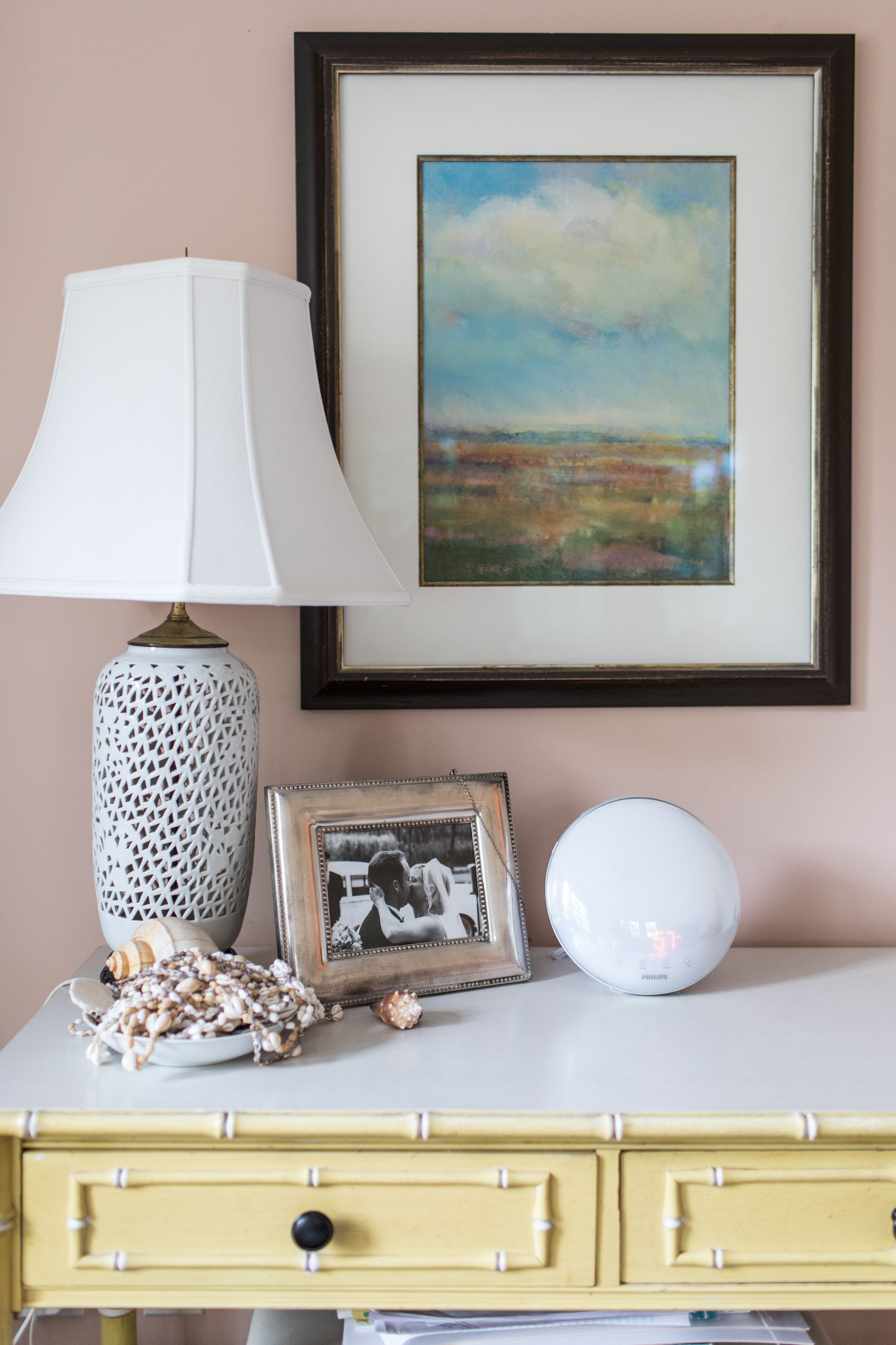 A yellow table holds a white lamp, framed landscape art, a black-and-white photo, seashells, a clock, and beads. Pink wall background.