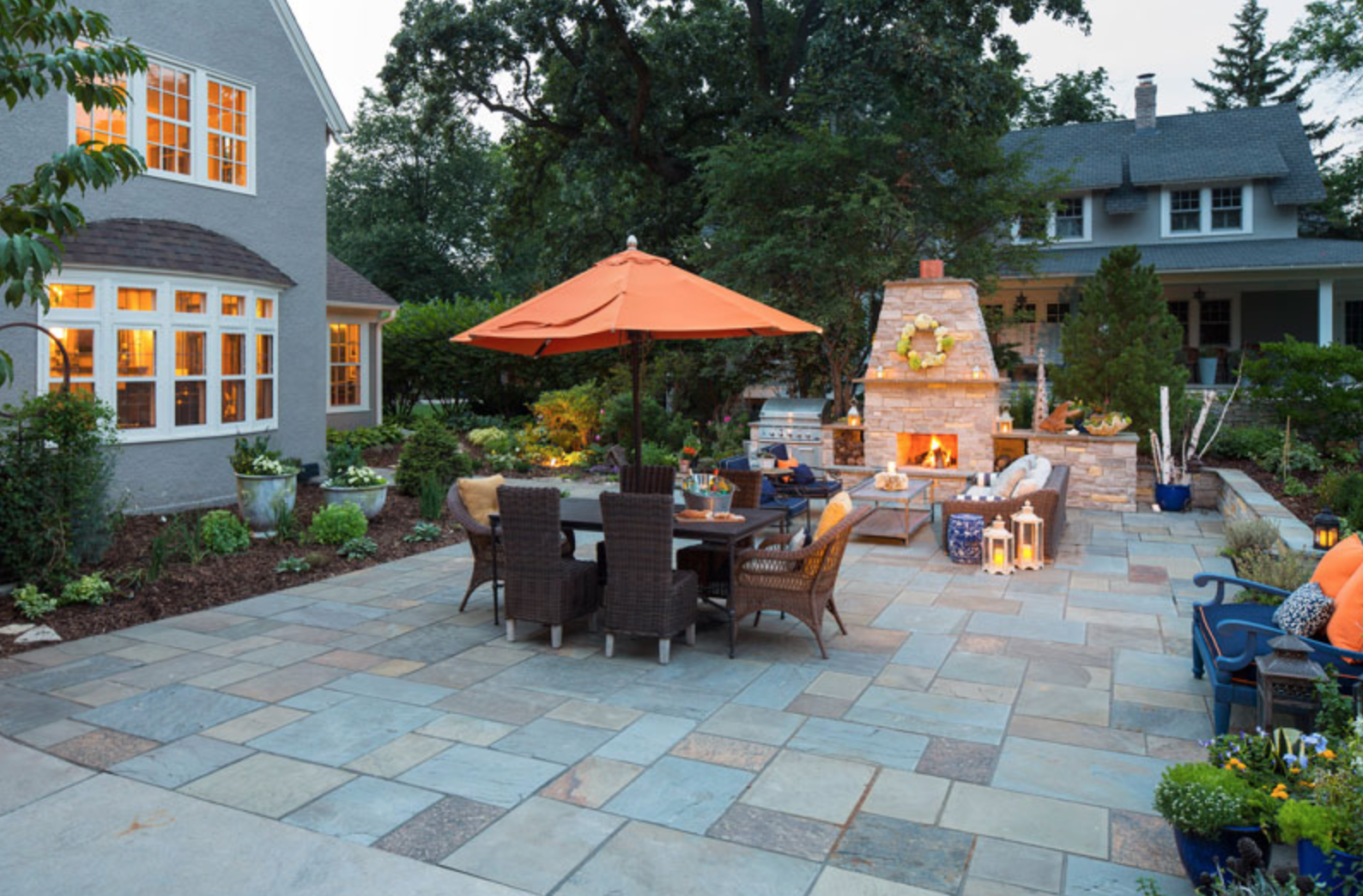 Patio with wicker chairs, orange umbrella, lit fireplace, and decor. Evening setting with warm lights and a cozy atmosphere.