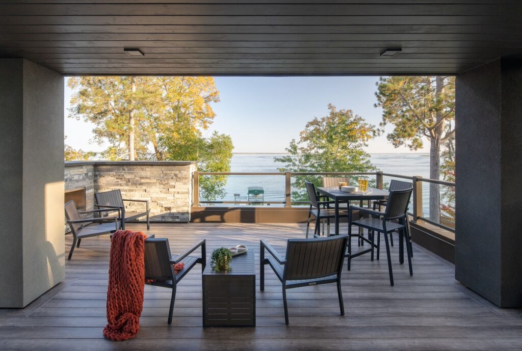 Modern patio overlooking a lake, featuring chairs, a table with drinks, a red blanket on a chair, green trees, and a serene, sunny view.