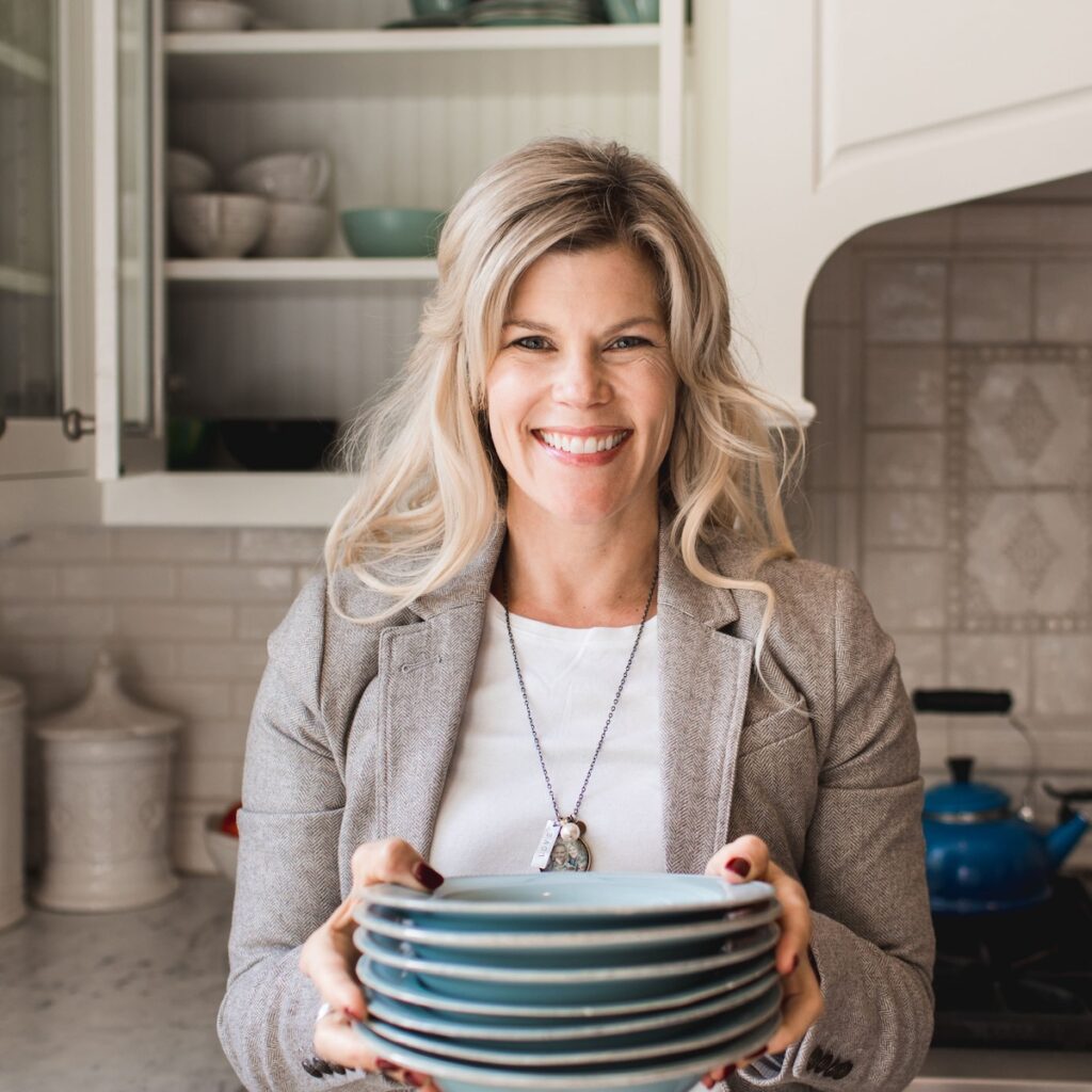 Smiling woman holding blue plates in a kitchen with open shelves, white cabinets, and a blue kettle, creating a warm, welcoming mood.