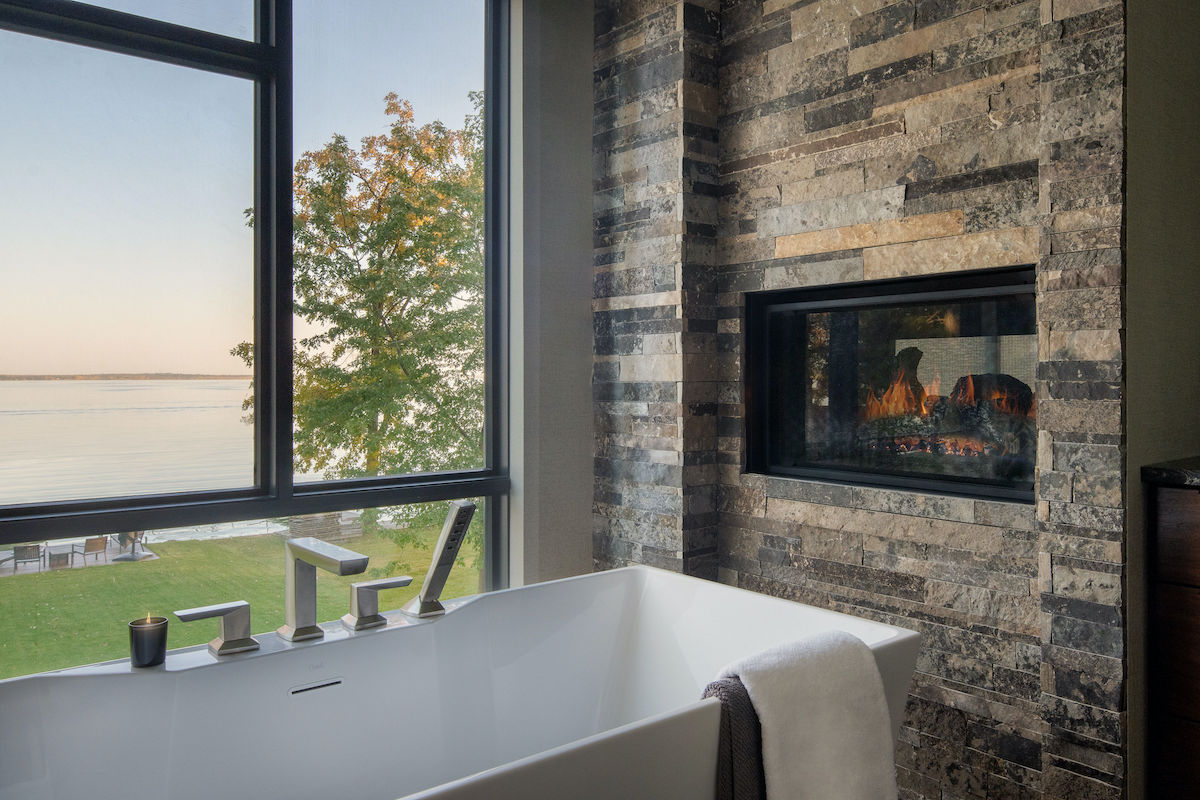 Modern bathroom with a white tub, gray towel, and lit candle by a large window showing a tree and lake view. Stone fireplace adds warmth.
