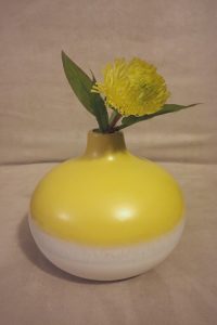 Yellow and white vase with a single yellow flower and green leaves, set against a plain beige background.